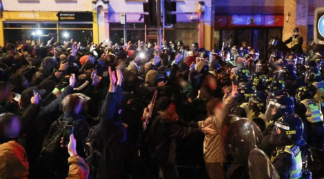 Lines of riot police backed by mounted units push into a crowd of protesters standing with their arms raised