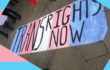 a banner painted blue pink and white in trans flag style, with black text reading TRANS RIGHTS NOW, it had been laid on the ground in front of some protesters, a raindbow gay pride flag can be seen flying behind it