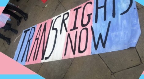 a banner painted blue pink and white in trans flag style, with black text reading TRANS RIGHTS NOW, it had been laid on the ground in front of some protesters, a raindbow gay pride flag can be seen flying behind it