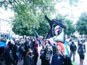 a crowd on college green, dozens of people with pride flags and anti homophia flags with trees in the background