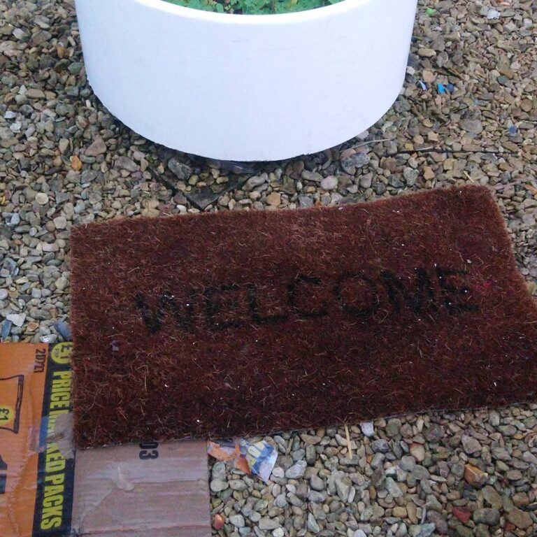 A worn Welcome mat that has been left on pebbles in a garden, with a potted plant next to it