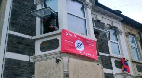 Two masked (and pixilated) people lean out of upstairs windows. Between them is a red flag with the squatting symbol that reads: Use it or loose it, squat.net