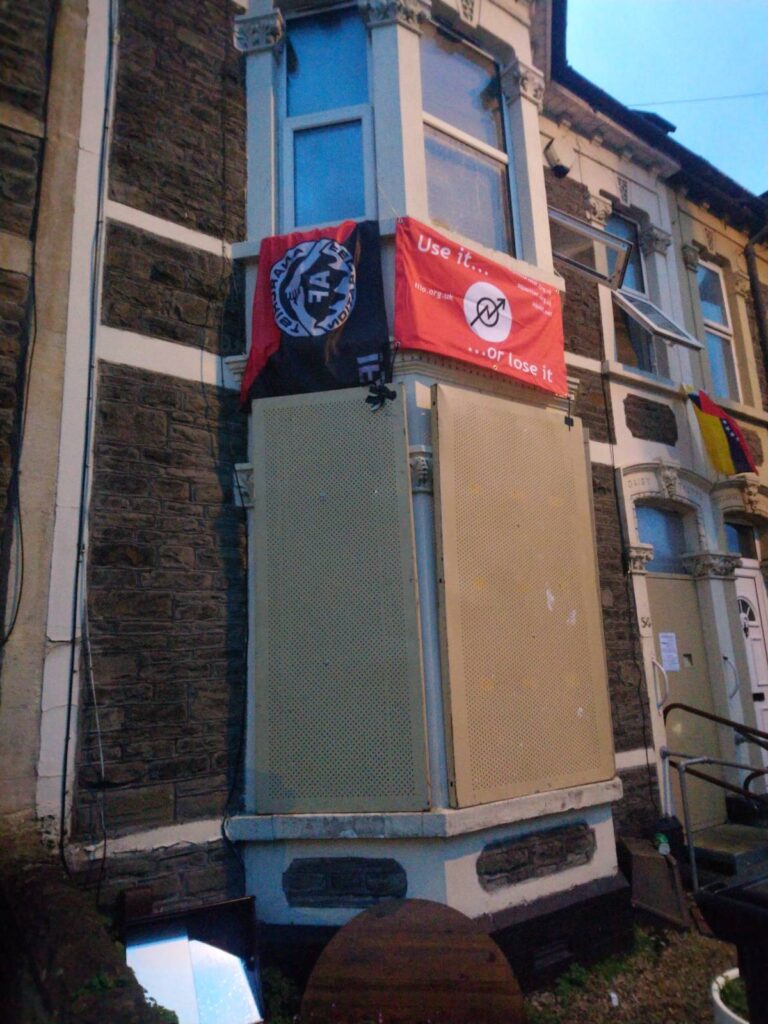a terraced house with metal citex boarding on the downstairs window and front door. flags hang from the top windows, including an Anarchist Federation Red and Black flag, and a squatting flag.