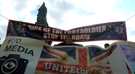 A fascist demonstrater lears down from between two banners one reads 'rise of the footsoldiers, stop the boats'